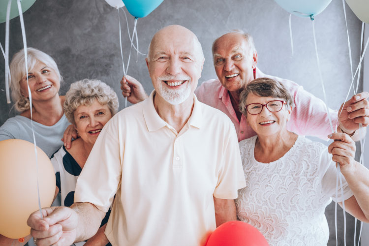 Four smiling mature men and women holding balloons gathered around a bald man with a short white beard celebrating a special occasion 