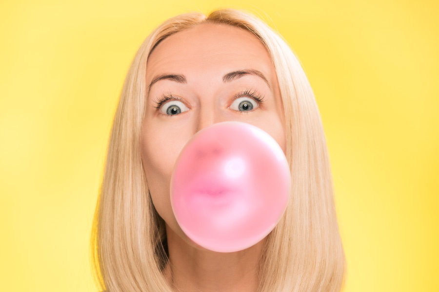 Blonde woman blowing a giant pink bubble gum bubble on a yellow background.